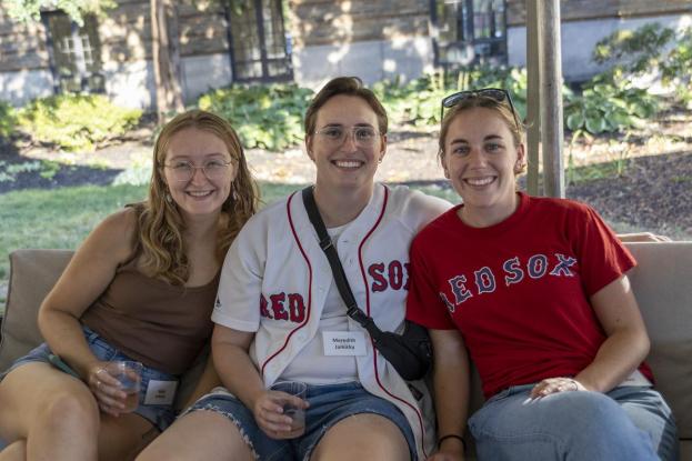 Three young alumni sit under a tent on the quad with their Red Sox jerseys on.