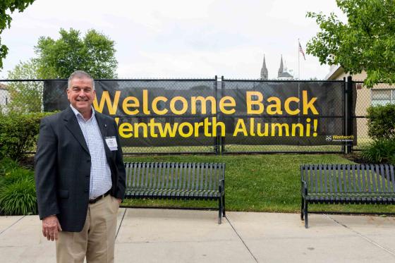 a man stands in front of a banner reading "Welcome Back Wentworth Alumni"